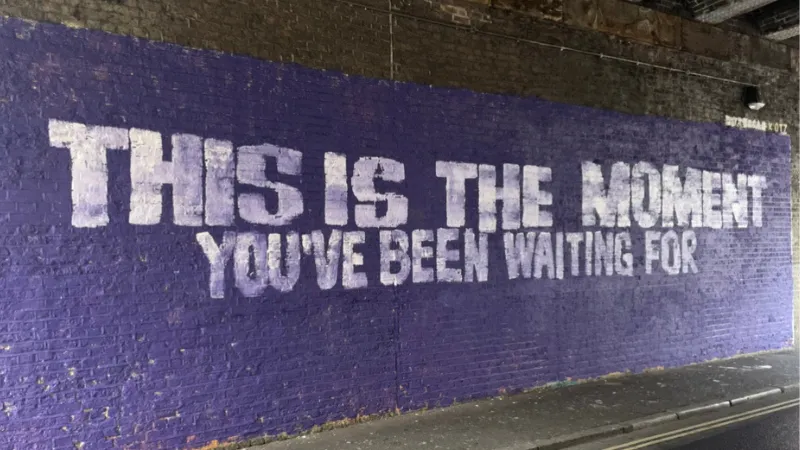 White graffiti text on a blue wall saying 'This is the moment you've been waiting for' in capital letters