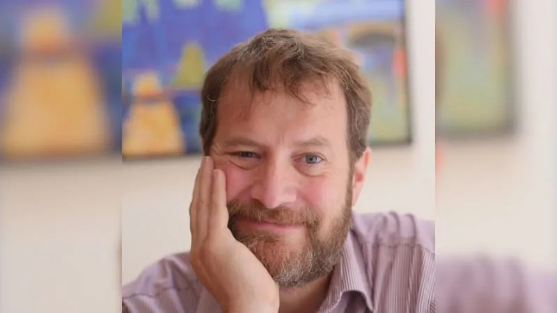 a bearded man wearing a striped shirt rests his chin on his right hand while smiling