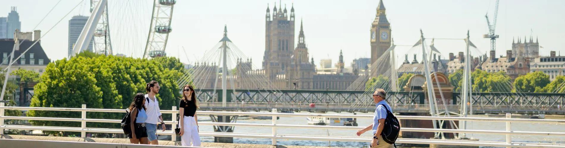 Three students walk along the Southbank along the River Thames with the Houses of parliament and a section of the London Eye in the background.