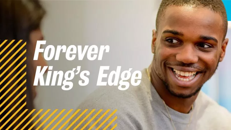 An image of a young Black man with short hair wearing a grey top, smiling while chatting to another person. The back of the head of the other person who has dark hair is in the leftmost part of the image. On the image is written Forever King's Edge