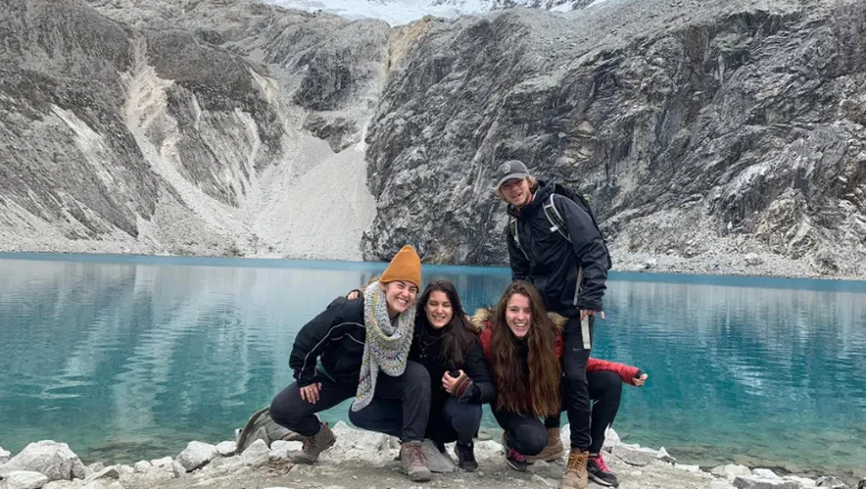 Four students dressed in winter clothes posing smiling in front of a lake and snowy mountains.