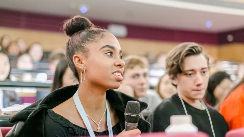 A close up of two students in an amphitheatre. The student on the left is a young woman holding a microphone. There are more students in the blurred background.