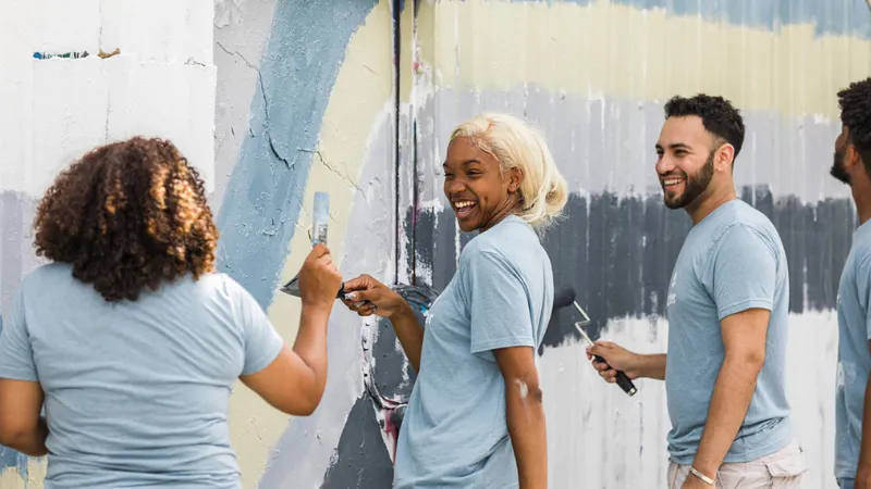 Four students wearing grey shirts, laughing and painting a wall.