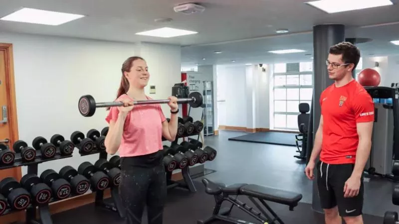 a woman in a gym lifts a barbell while being spotted by a personal trainer