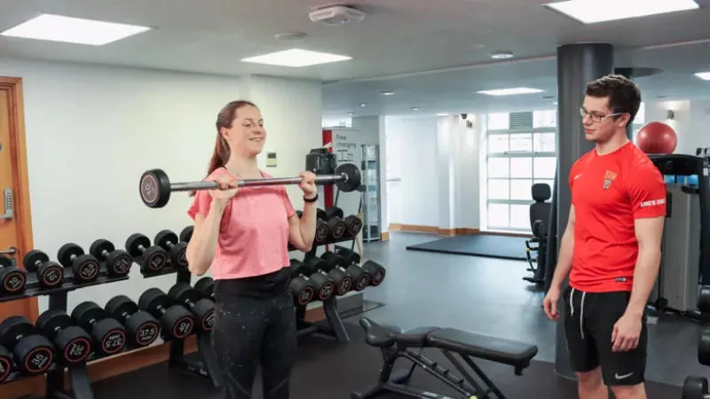 a woman in a gym lifts a barbell while being spotted by a personal trainer