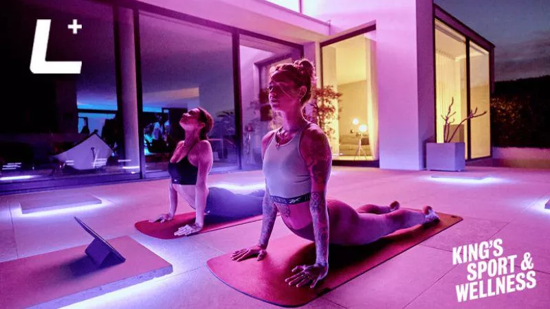 Two women do Yoga in an apartment with purple lighting, the Les Mills + logo and King's Sport and Wellness logo in the left corner.