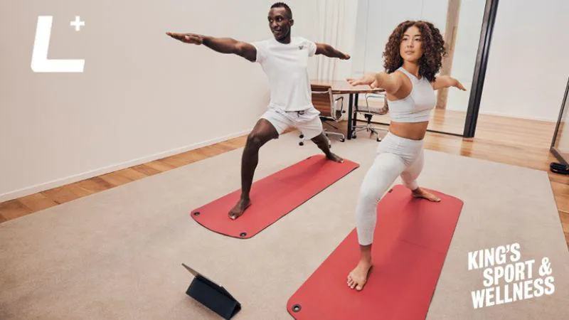 A man and a woman doing a Yoga class in their own home with the Les Mills + logo and King's Sport and Wellness logo in the left corner.