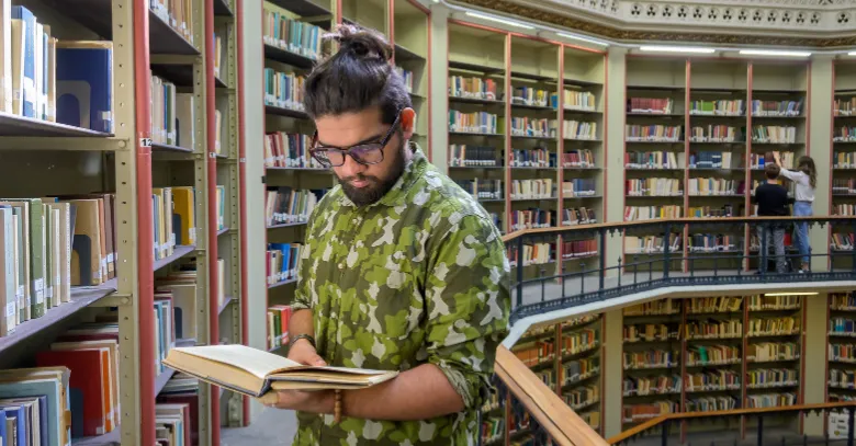 A student stood in front of a row of bookshelves reading a book in King's Maughan Library