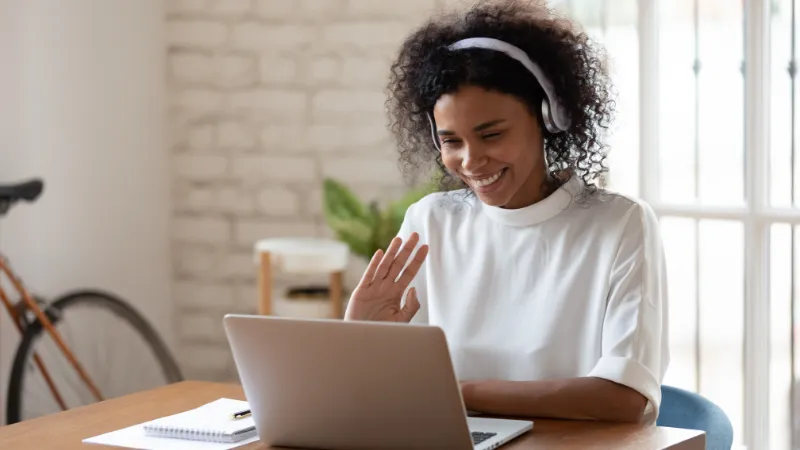 A Black female student wearing headphones, sitting at a table in front of an open laptop being on a call and smiling.