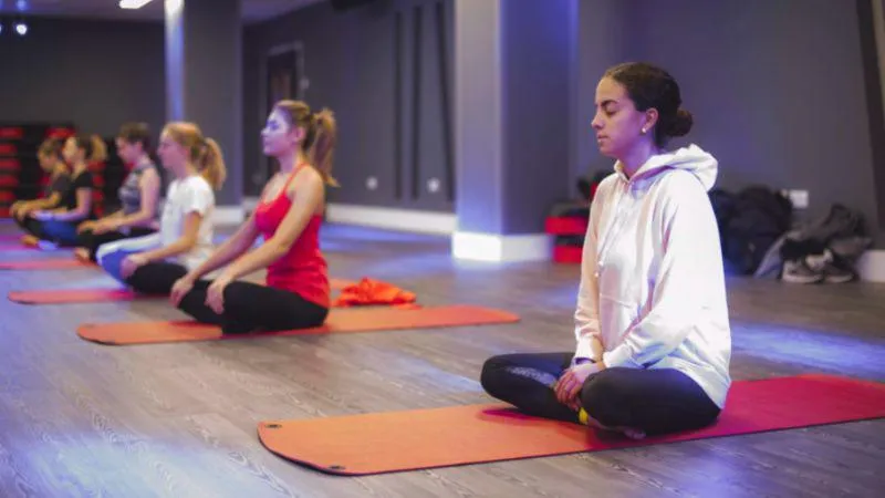 Image shows an exercise studio with wooden flooring and grey walls, a woman in a white hoodie sits on a red yoga mat, with other people taking place in the class in a line to the left.