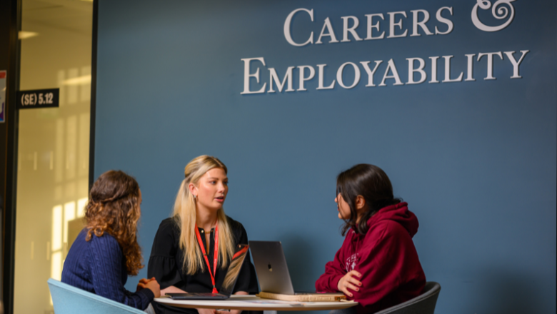 Image of two students sat at a table speaking to a member of King's staff. A blue wall is behind them with the words: Careers & Employability
