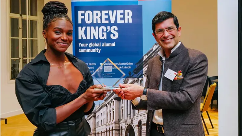 An image of Dina Asher-Smith receiving a King's Distinguished Alumni award from President & Principal Shitij Kapur. They stand in front of a blue banner that says Forever King's and are holding a glass award trophy.