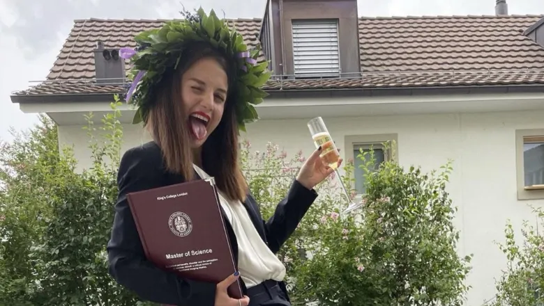 A student wearing a laurel wreath on their head, holding their bound thesis and a drink n the other hand