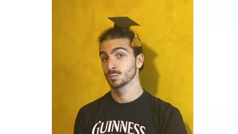 A student wears a small cardboard mortarboard against a yellow wall