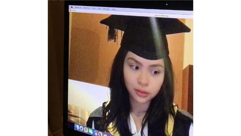 Student looking at themselves in a computer screen wearing a graduation mortarboard and gown