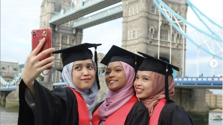 Three students taking a selfie in front of Tower Bridge wearing graduation mortarboards and gowns with red stoles
