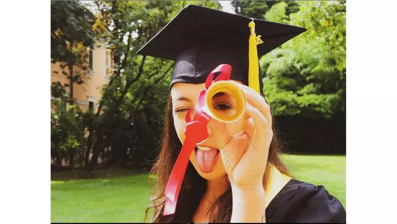 A student wearing a mortarboard sticking their tongue out and holding a rolled up scroll to their left eye