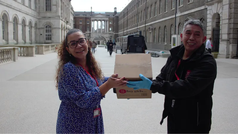 An image of a woman and a man in the Strand Campus courtyard passing a Hey Girls cardboard box from the first delivery.