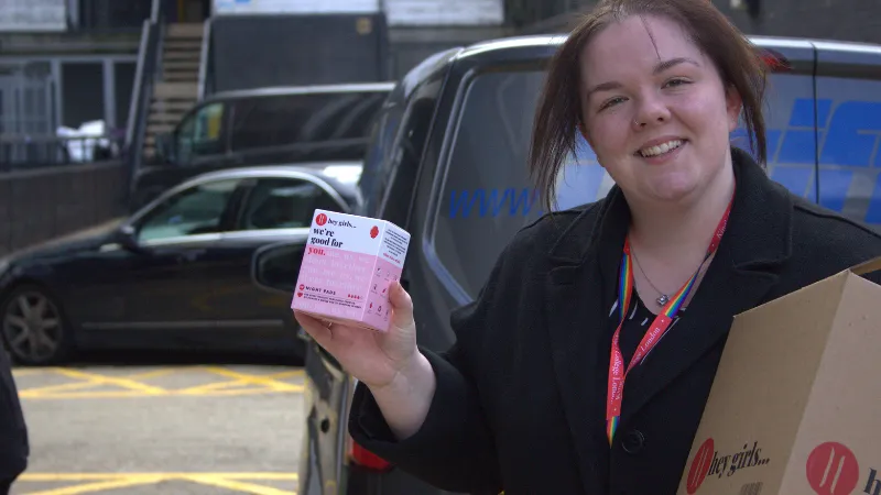 A member of King's Staff unpacking a delivery from Hey Girls, the woman in the image is wearing a black King's zip up jacket with a pink and white Hey Girls product box.