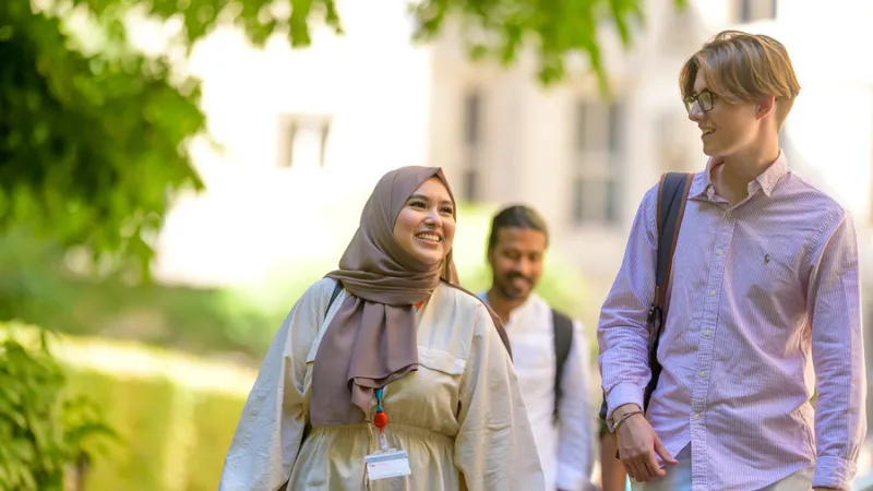 An image of 3 students walking, chatting and smiling in front of a background of a building and tree. One student is a young woman wearing a head scarf, another is a young man with blond hair and the third is a young man with dark hair.