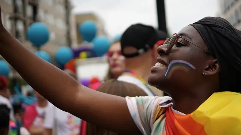 A Black person with a rainbow drawn on their face participating in Pride with more people in the background.