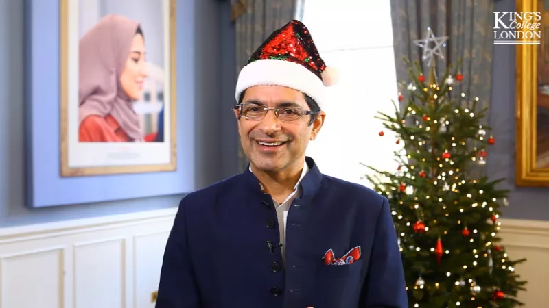A man wearing a blue suit and sparkly Santa hat stands in front of a Christmas tree covered in lights and baubles