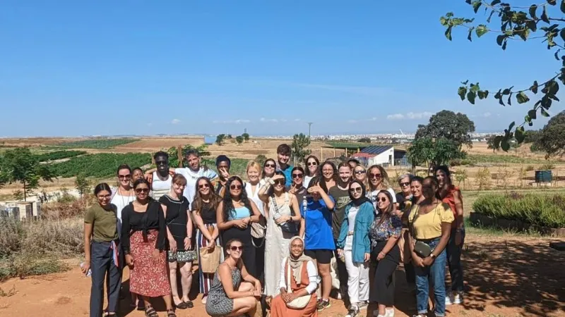 A group of students posing in a sunny Mediterranean landscape with low vegetation and a city in the far background.