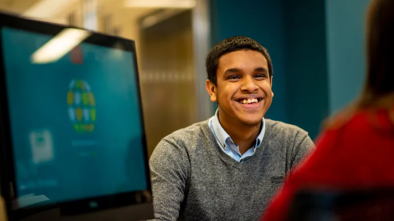 Student smiling at another person sat at a desk with a computer