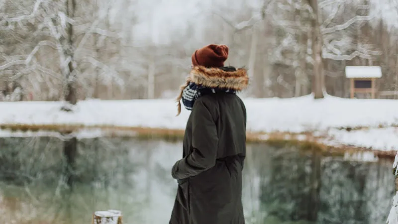 a figure wearing a thick green coat and red woollen hat stands in front of a small lake in a snowy forest