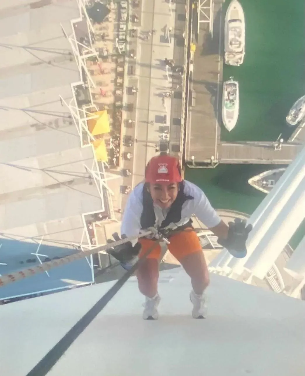 a young person photographed from above as they abseil down a tower. they are wearing a red hard helmet and attached to a rope and harness. boats, jetties and water can be seen below them.