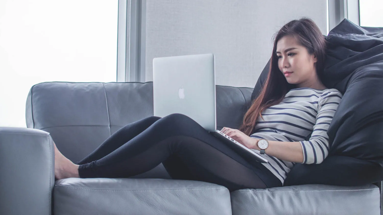 a student lies on a grey sofa while using a laptop