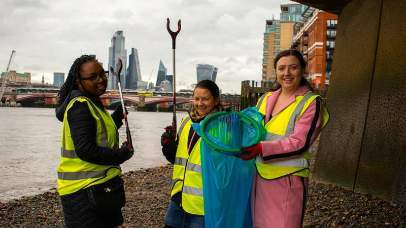 Three students stand on the banks of the river Thames, they are collecting rubbish