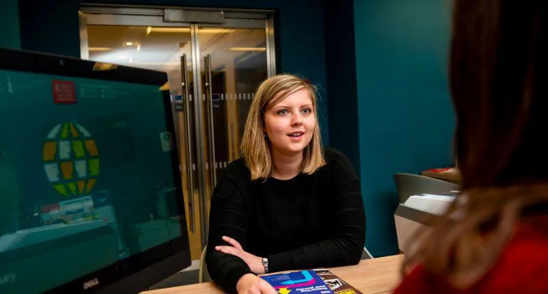 Image of a student sat opposite a desk and talking to someone