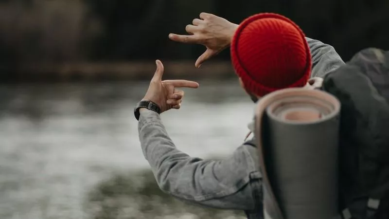 A hiker wearing a backpack and red woolen hat uses their fingers to create a frame around a view of a lake