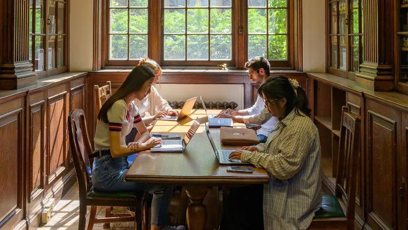 Four students sit at a wooden table in the library, each student has a laptop in front of them.