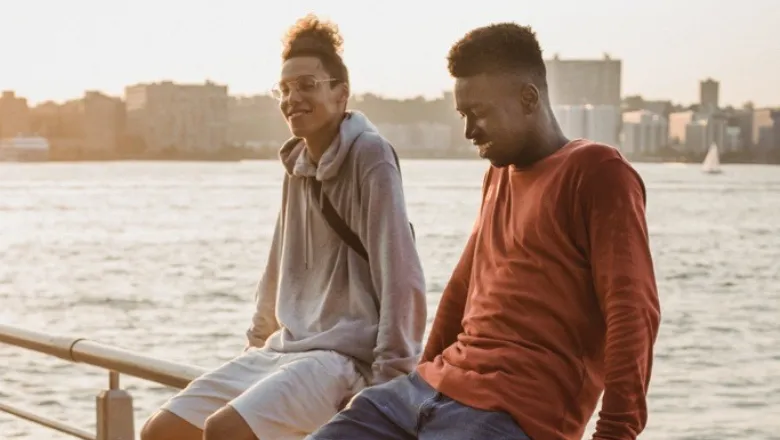 Two people sat on a railing next to a river smiling and laughing