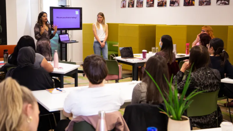 A woman presenting with a TV screen to a room of people sat at tables
