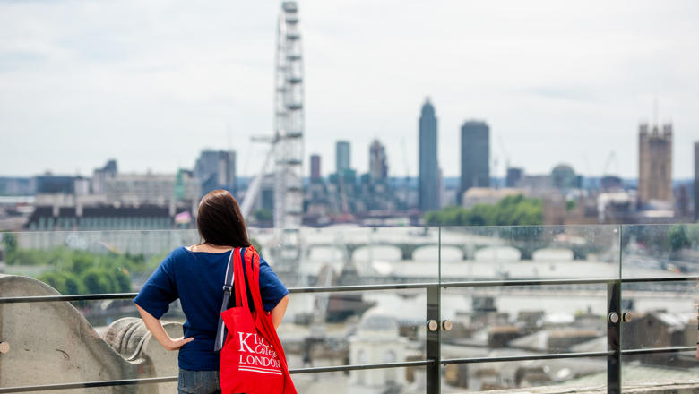 Woman looks out over London skyline with red King's College London bag on shoulder.
