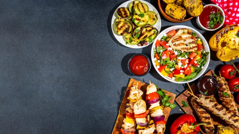 Assorted grilled vegetables, meat skewers, grilled chicken salad, corn on the cob, and dipping sauces arranged on a dark table.