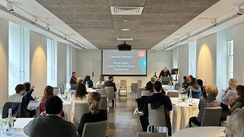 The audience sits at tables listening to a speaker presenting at the front of the room.